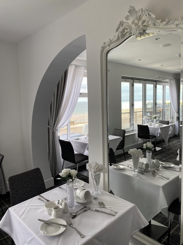 A dining area with tables set for service, a large mirror reflecting the room, and a view of the sea through large windows.