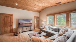 A spacious living room with a wooden ceiling, a large flat-screen TV, a sofa, and a window with a view of trees. - Monthly Lease - Freedom Lodge by Tahoe Getaways - 4BR Centrally Located w/ Bonus Room (Truckee)