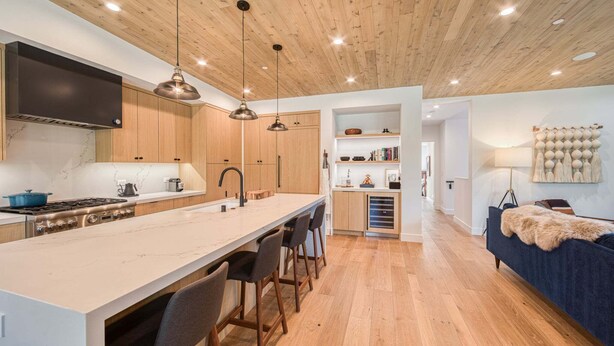 Een moderne keuken met houten kasten, een centraal eiland, hangende lampen en uitzicht op de woonkamer.