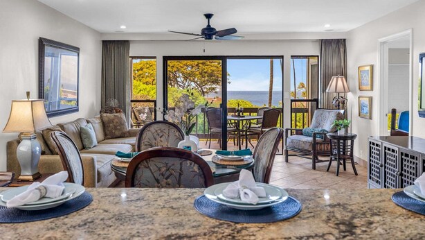 A spacious room with a dining area, a sofa, a ceiling fan, and large windows with a view of the ocean. - Sunlit 2BR Island Haven Plantation Style (Lahaina)