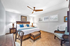 Un dormitorio con cama, mesitas de noche, una silla, una mesa, ventilador de techo y ventana con persianas.