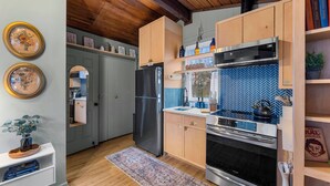 A kitchen with wooden cabinets, a stainless steel refrigerator, a microwave, and an oven. There is a blue herringbone backsplash and a wooden ceiling. - The Nook - Cozy Bungalow in the Countryside (Weaverville)
