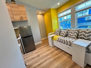 A compact living space with a kitchenette, a sofa with patterned cushions, and a window with a view of a blue building.
