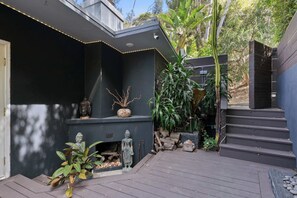 A modern outdoor area with a dark-colored wall, a Buddha statue, and a staircase leading to a higher level. - Hollywood Hills Vista | Decks & Fire Pit by C&C (Los Angeles)