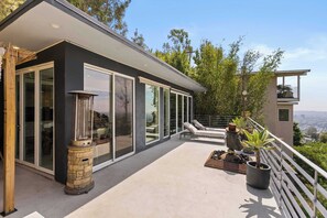 A modern house with large glass windows, a stone fireplace, outdoor seating, and a view of the city.