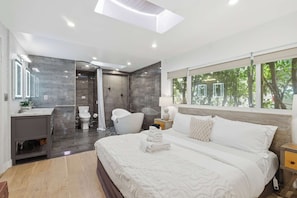 Un dormitorio moderno con una cama grande, una mesita de noche, un tocador, una ventana con vistas a árboles y un baño con bañera e inodoro.