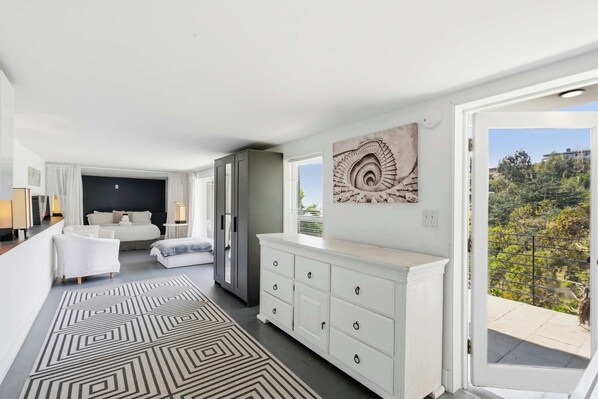 A modern bedroom with a bed, a dresser, a painting, and a view of greenery.