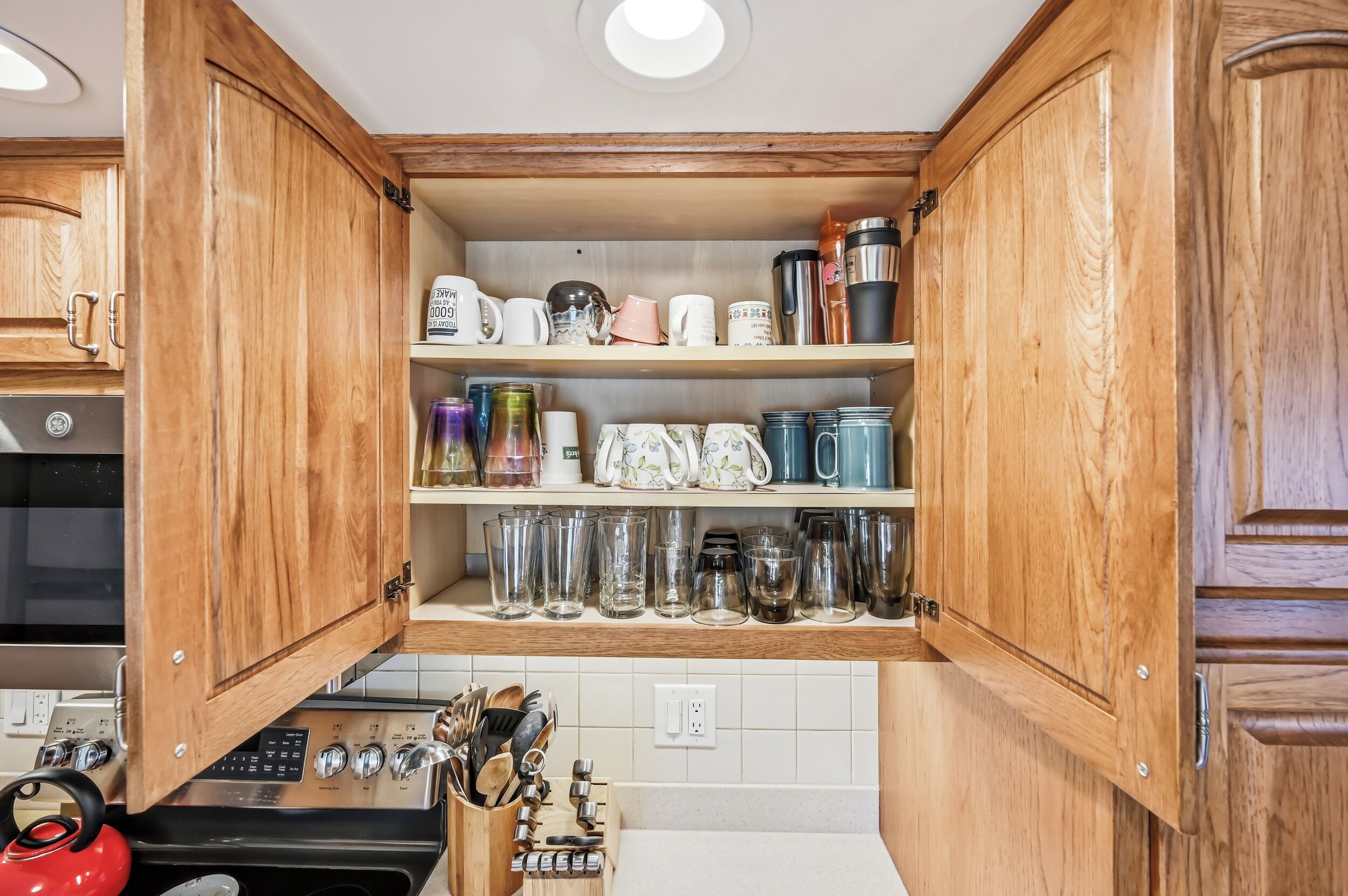 Una cocina con armarios de madera, estufa y diversos utensilios de cocina, incluyendo tazas, vasos y cristalería.