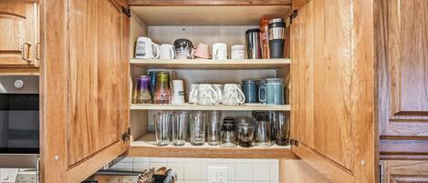Una cocina con armarios de madera, estufa y diversos utensilios de cocina, incluyendo tazas, vasos y cristalería.
