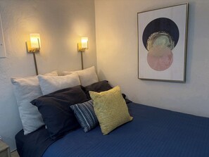 Un lit avec plusieurs oreillers, deux lampes fixées au mur et une œuvre d’art abstraite encadrée accrochée au mur.