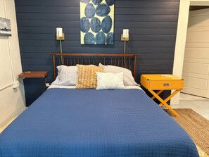 Un lit avec une couette bleue, une tête de lit en bois et plusieurs oreillers. On y trouve une table de chevet avec une lampe, une petite table avec une télécommande et un luminaire fixé au mur.