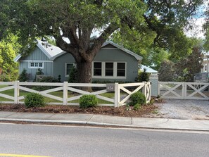 Une maison à deux étages, avec un grand arbre devant, une clôture blanche en bois et une allée menant à une entrée fermée par un portail.