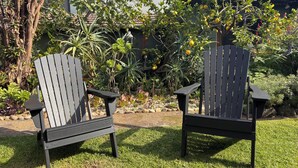 Two Adirondack chairs in a garden with various plants and a building in the background. - Retro bungalow in organic garden oasis (Caulfield North)