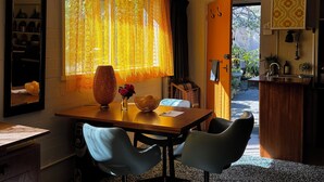 A dining area with a wooden table, two chairs, a vase with flowers, and a window with yellow curtains. - Retro bungalow in organic garden oasis (Caulfield North)