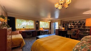 A hotel room with a large bed, a desk, a chair, a small kitchenette, and a bookshelf. - Retro bungalow in organic garden oasis (Caulfield North)