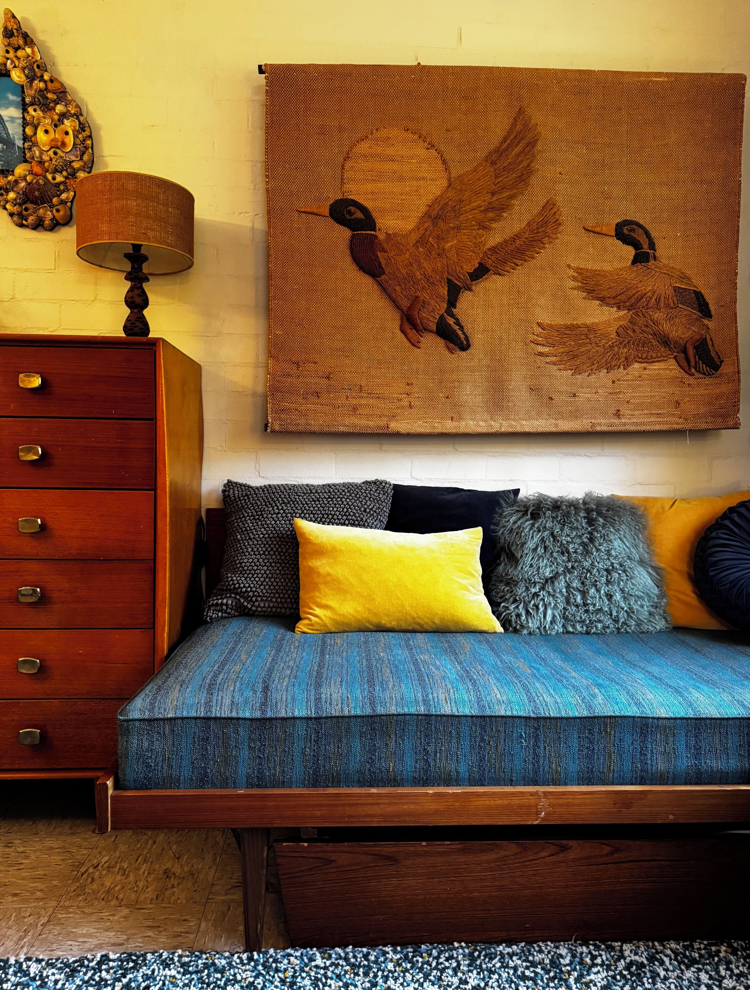 A bed with a blue and yellow bedspread, a wooden dresser with drawers, a lamp, and a wall-mounted tapestry.