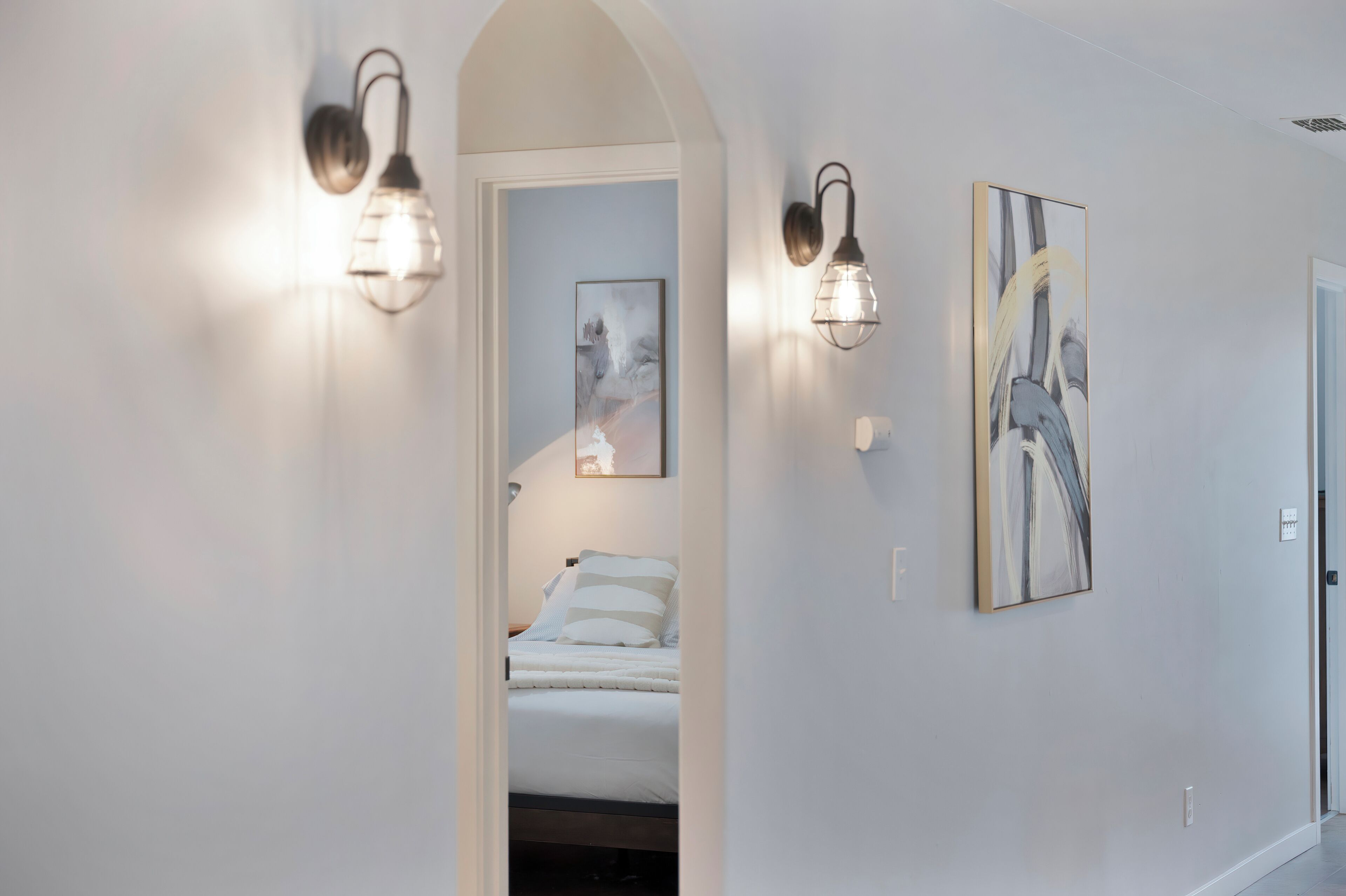 A hallway with a bed in the background, two wall-mounted light fixtures, and two framed artworks on the wall.