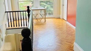 Una stanza con pavimento in legno a spina di pesce, ampie finestre, una scala e una porta rossa.