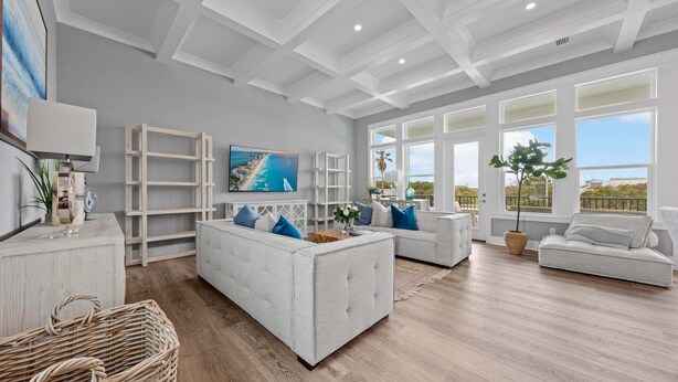 Un salón amplio con un televisor grande, sofás blancos, suelo de madera y ventanas grandes con vistas.