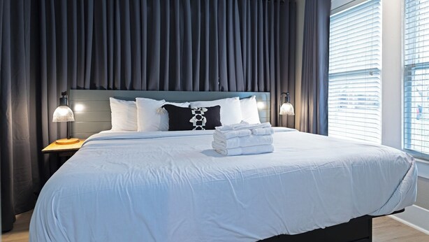 A neatly made bed with white linens, a headboard with a geometric pattern, and a bedside lamp. - 2BR Lower Westheimer Dream 01! Walkable to Montrose! (Houston)