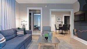 A modern living room with a grey sofa, a coffee table with decorative items, a dining area with a table and chairs, and a large mirror reflecting the room. - 2BR Lower Westheimer Dream 01! Walkable to Montrose! (Houston)