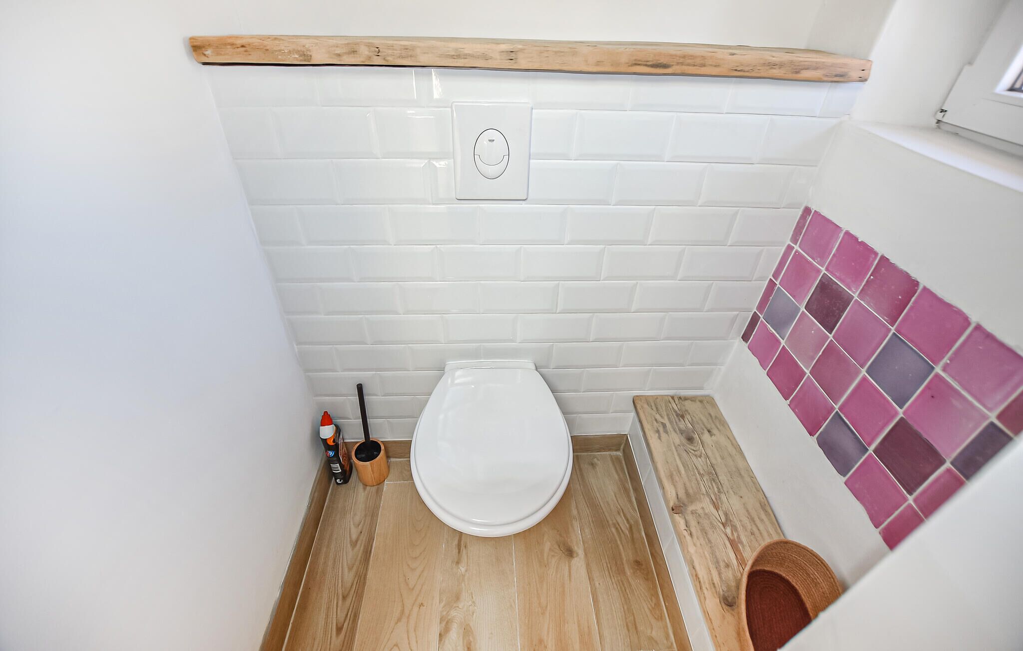 A small bathroom with a white toilet, wooden floor, white tiled walls, and a pink-tiled shower area.
