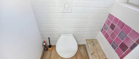 A small bathroom with a white toilet, wooden floor, white tiled walls, and a pink-tiled shower area.