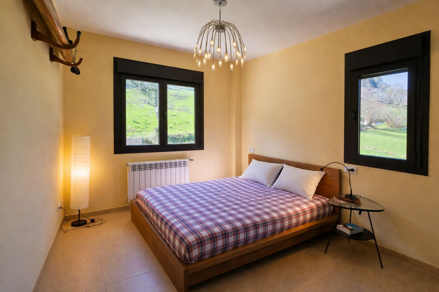 A bedroom with a bed, a bedside table, a lamp, a radiator, and two windows with a view of greenery.