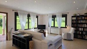 En moderne stue med sofa, fjernsyn, bogreol og store vinduer med gardiner.