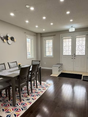 Un espace repas comprenant une table, des chaises et un tapis à motifs. On y trouve deux fenêtres équipées de stores, une porte blanche avec un panneau en verre et un luminaire au plafond.
