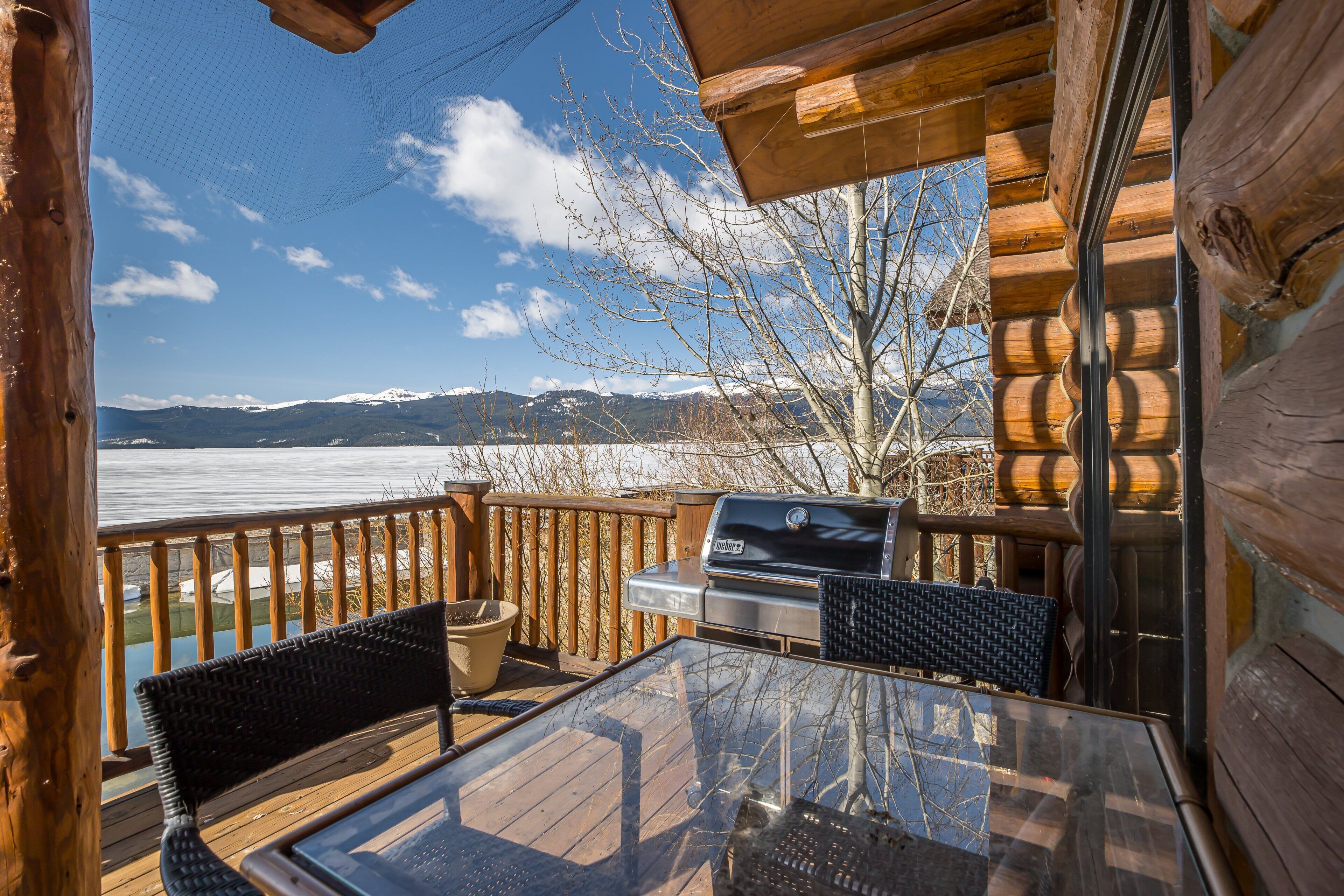 A wooden deck with a grill and chairs, overlooking a body of water and mountains.