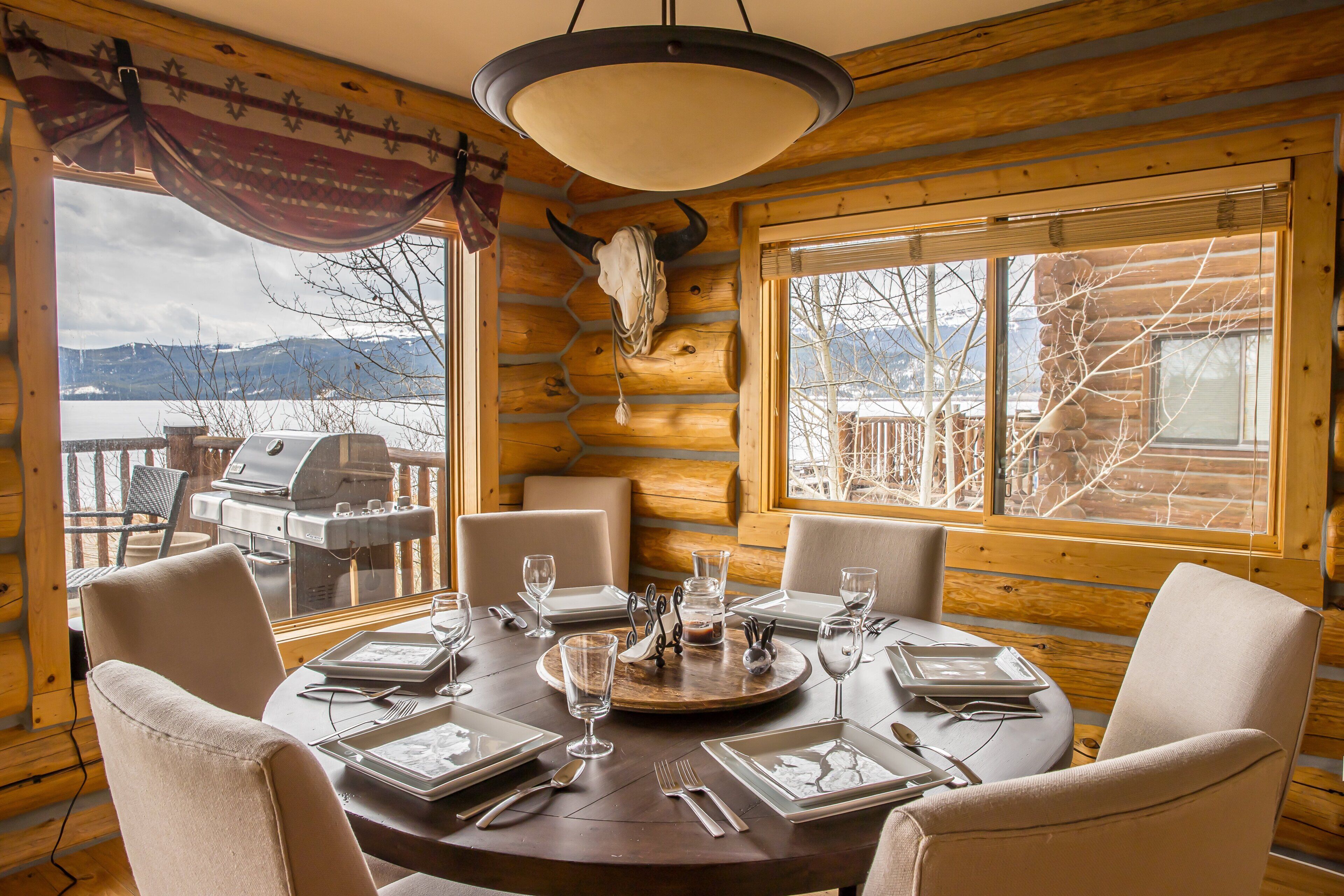 A log cabin dining room with a round table set for six, a large window with a mountain view, and a wooden ceiling with a cow skull decoration.