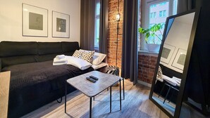 Ein modernes Zimmer mit Backsteinwand, einem schwarzen Sofa, einem kleinen Tisch mit Fernbedienungen und gerahmten Kunstwerken an der Wand.