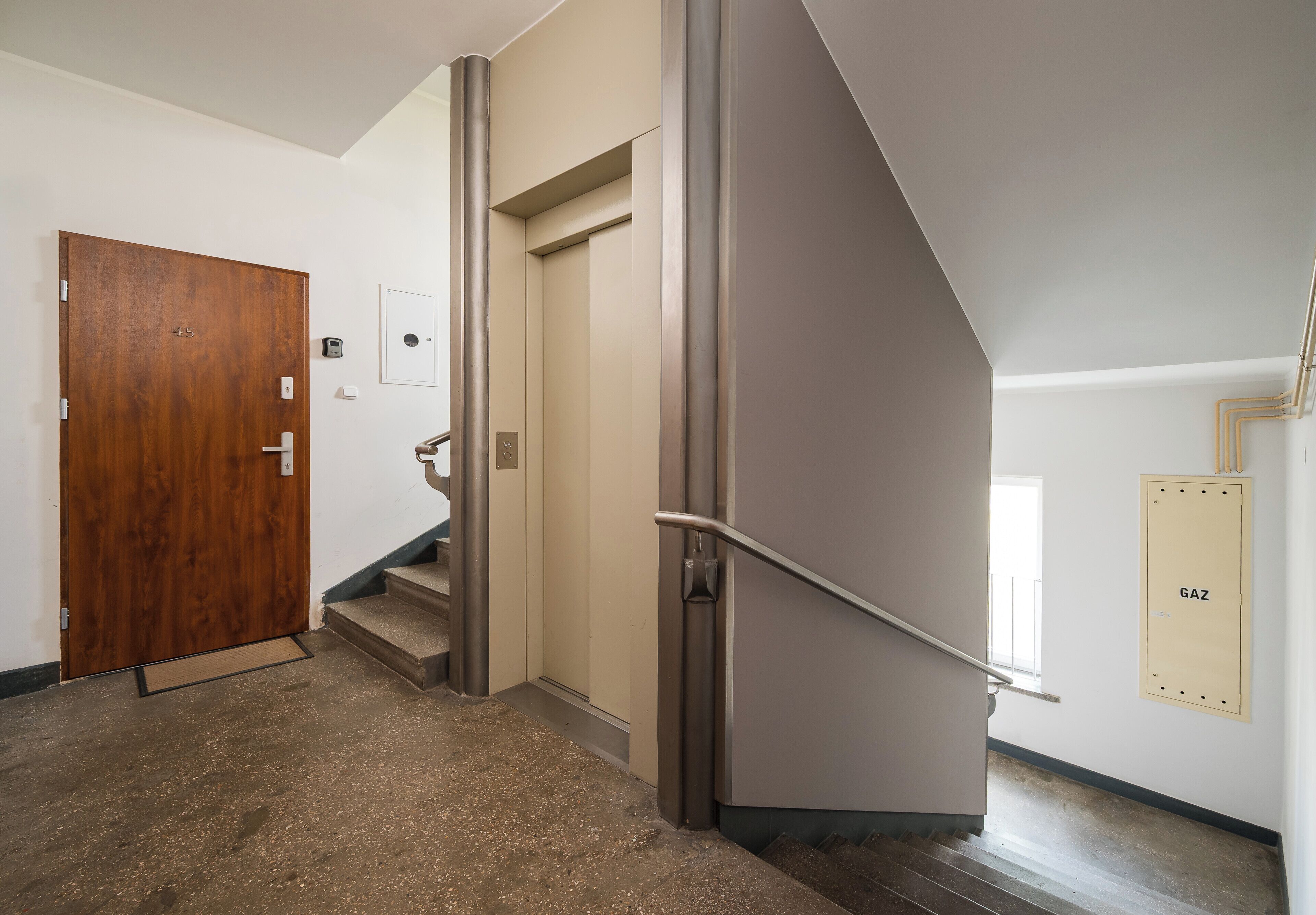 Uma escada com corrimão de metal, porta de madeira e quadro elétrico fixado na parede com a etiqueta 'GAZ'.