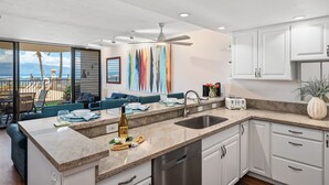 A modern kitchen with a large island, a built-in dishwasher, and a view to a balcony. - SUMMER SALE! Remodeled Oceanfront 1BR Steps to Pool & Ocean - Kahana Reef 107 (Lahaina)