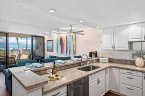 A modern kitchen with a large island, a built-in dishwasher, and a view to a balcony. - SUMMER SALE! Remodeled Oceanfront 1BR Steps to Pool & Ocean - Kahana Reef 107 (Lahaina)