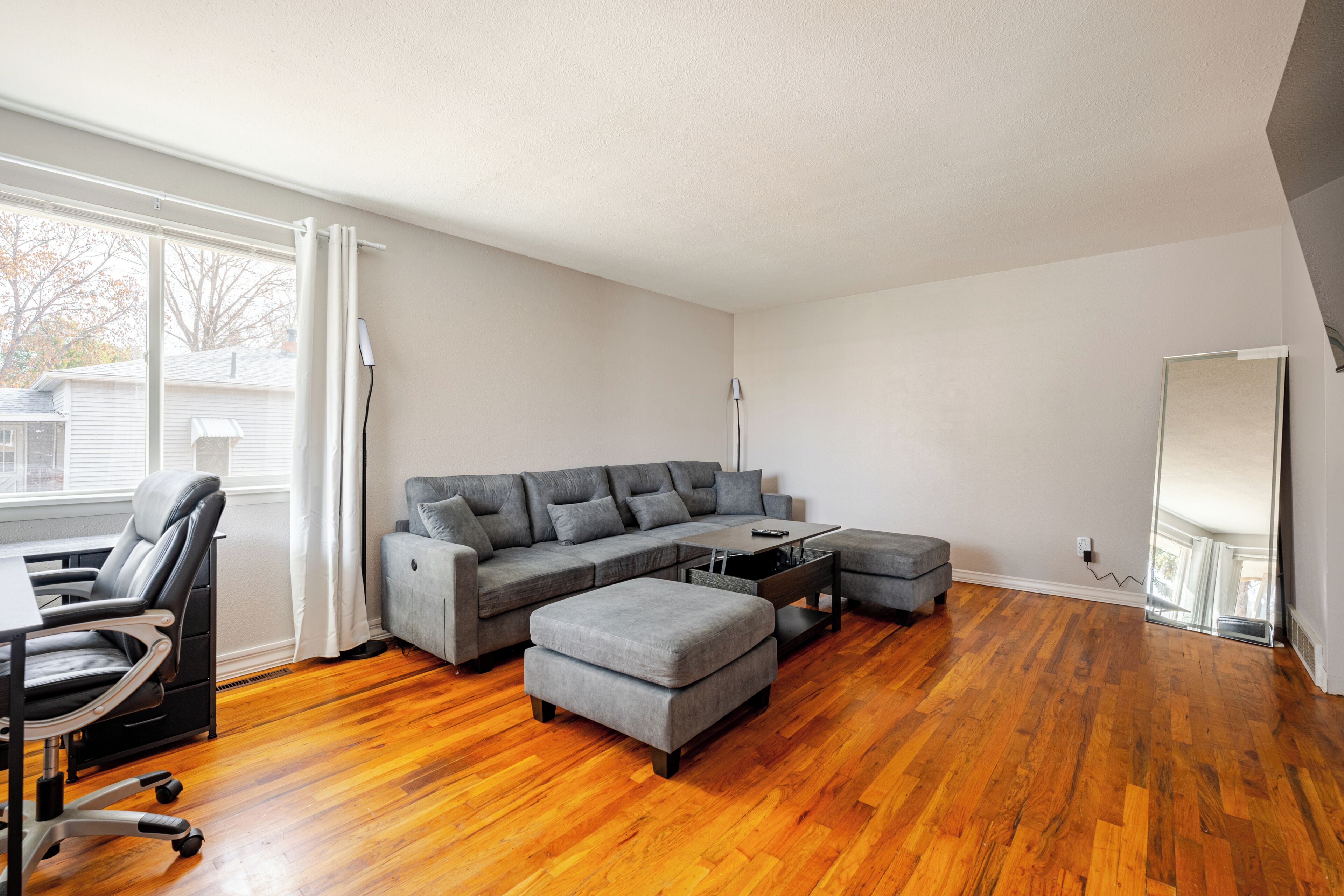 A modern living room with hardwood flooring, a sectional sofa, a coffee table, and a desk with a chair.