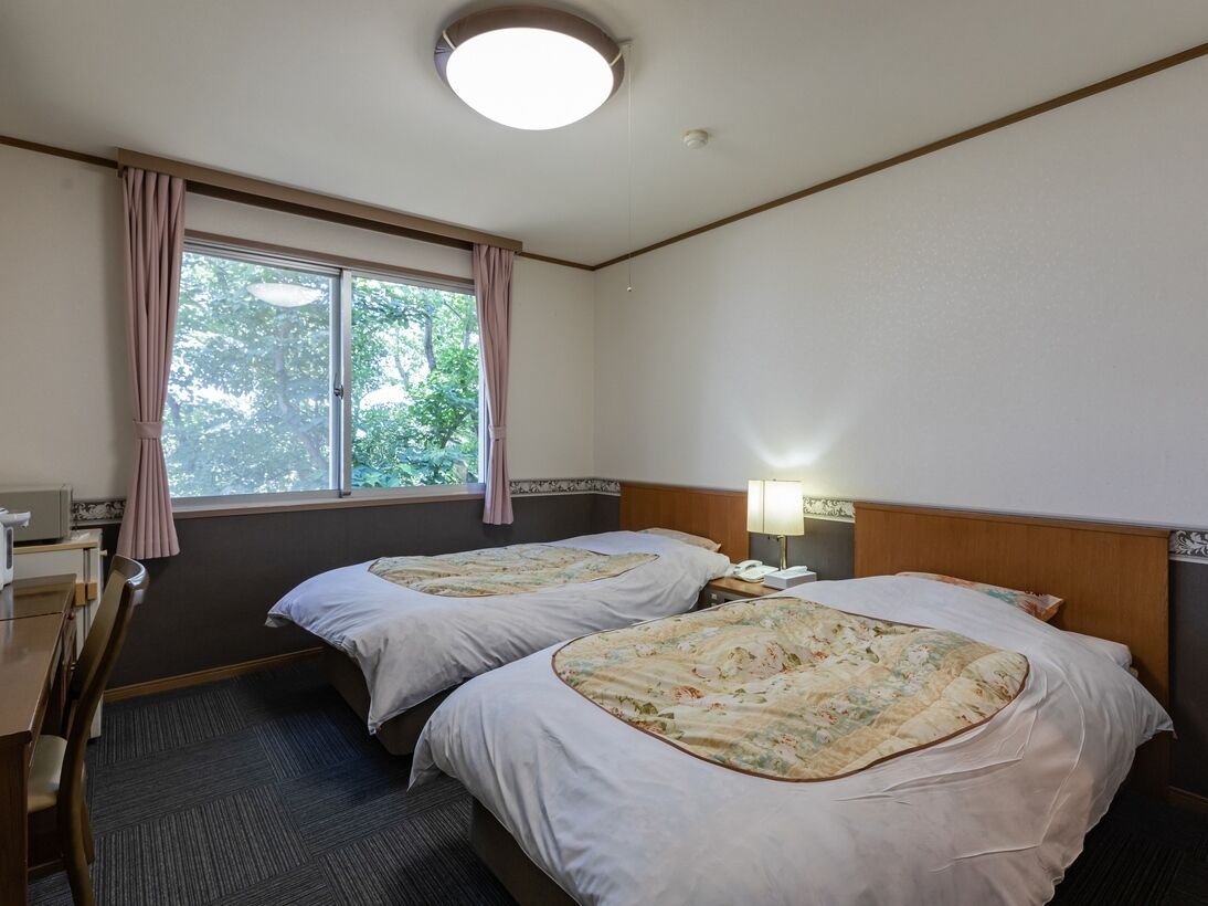 A hotel room with two beds, a desk, a chair, a lamp, and a window with curtains.