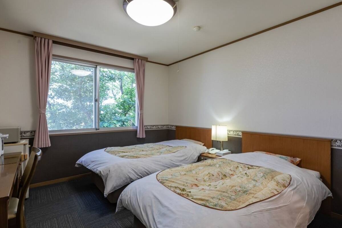 A hotel room with two beds, a desk, a chair, a lamp, and a window with curtains.