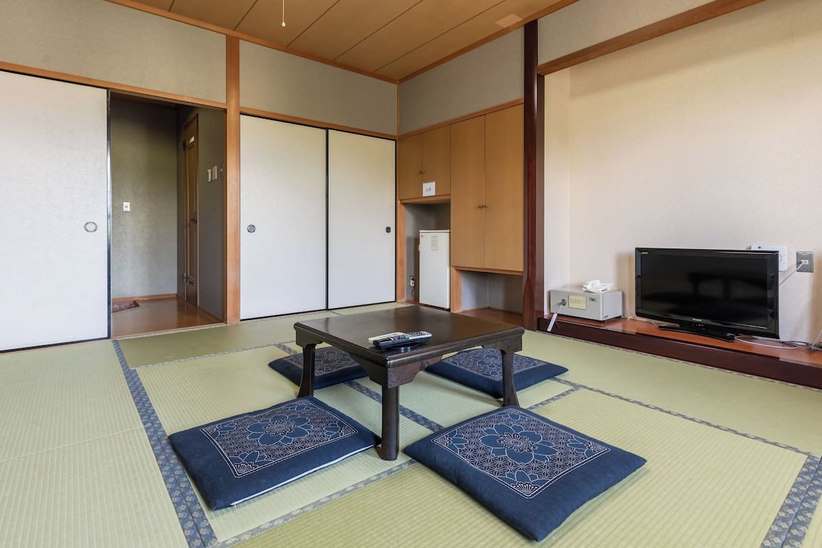 A traditional Japanese room with tatami flooring, a low table, cushions, a television, and sliding doors.