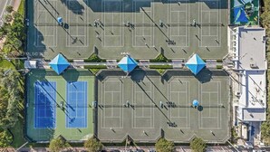 Vista aérea de un complejo de canchas deportivas con canchas de tenis y una cancha de baloncesto, rodeado de edificios y estacionamiento.