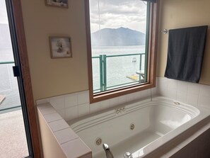 A bathroom with a bathtub, a window with a view of water and mountains, a towel rack, and a framed picture on the wall.