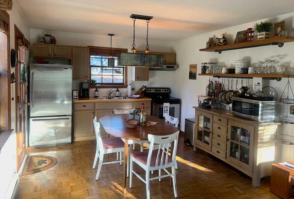 A kitchen with wooden cabinets, a stainless steel refrigerator, a wooden dining table with chairs, and a hanging light fixture.
