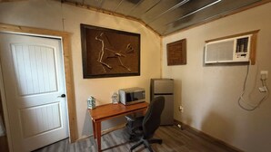 A room with a wooden desk, a chair, a white door, a framed artwork, a refrigerator, and an air conditioning unit.