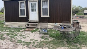 A small, single-story house with a front door, two windows, a porch, and a patio with metal chairs and a table.