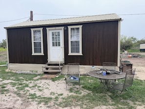 A small, single-story house with a front door, two windows, a porch, and a patio with metal chairs and a table.