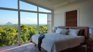 Ein Schlafzimmer mit großem Fenster, einem Bett, einem Nachttisch und Blick auf einen Wald.