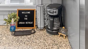 Uma cozinha com um armário que contém copos, uma cafeteira e uma placa com a inscrição 'WELCOME TO THE URBAN RETREAT'.
