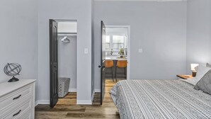 Quarto com cama, penteadeira, escrivaninha e cadeira. Há uma porta que dá para um banheiro e uma cozinha.