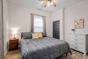 Un dormitorio con cama, mesita de noche, cómoda, ventana con cortinas y ventilador de techo.